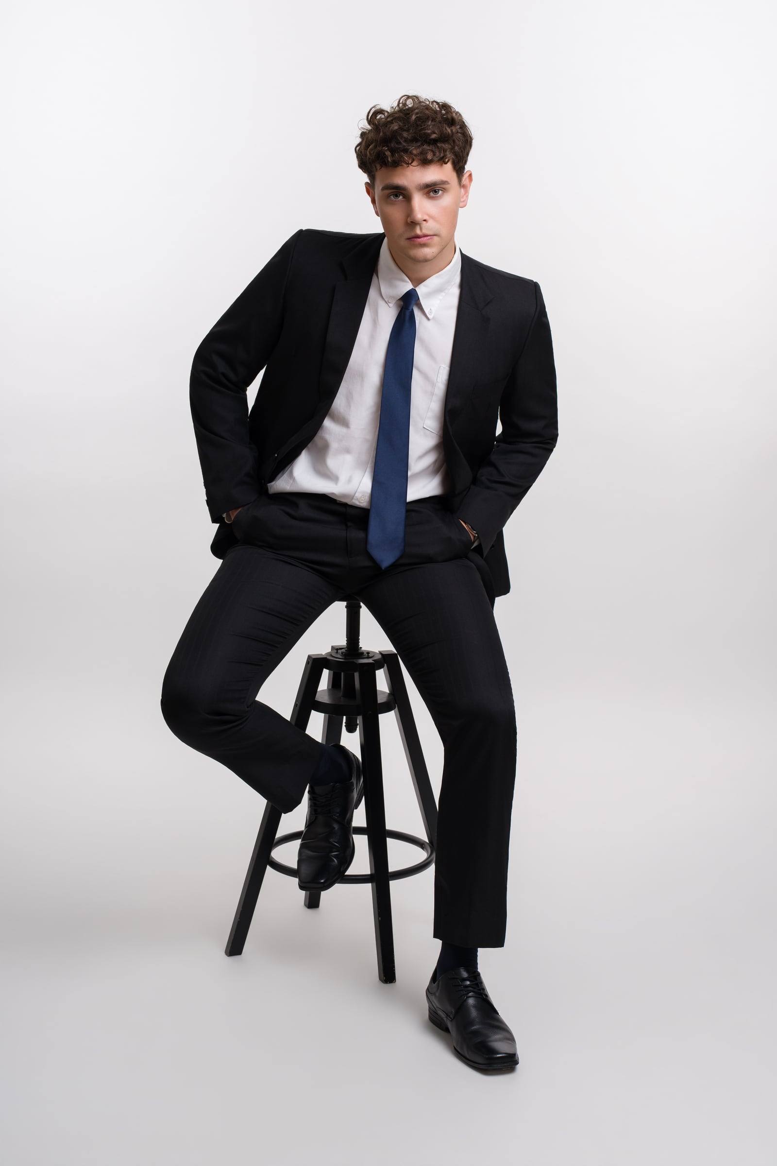 Jorge Tyrakowski in a black suit, white shirt and navy tie, seated on a stool