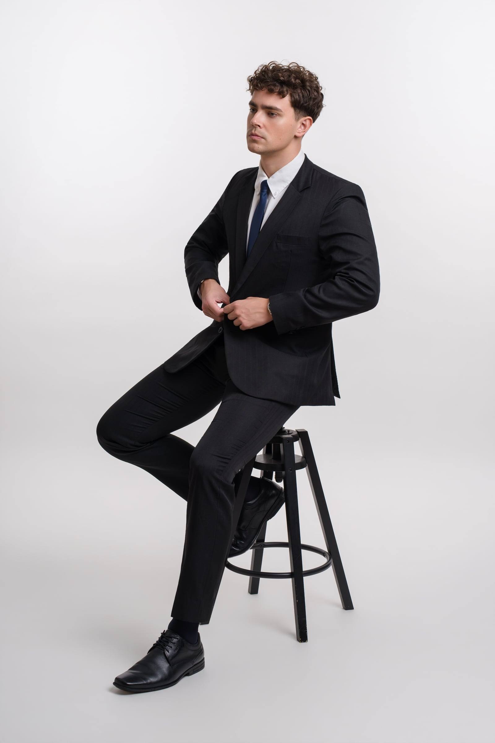 Jorge Tyrakowski in a black suit, white shirt and navy tie, seated on a stool, buttoning the jacket